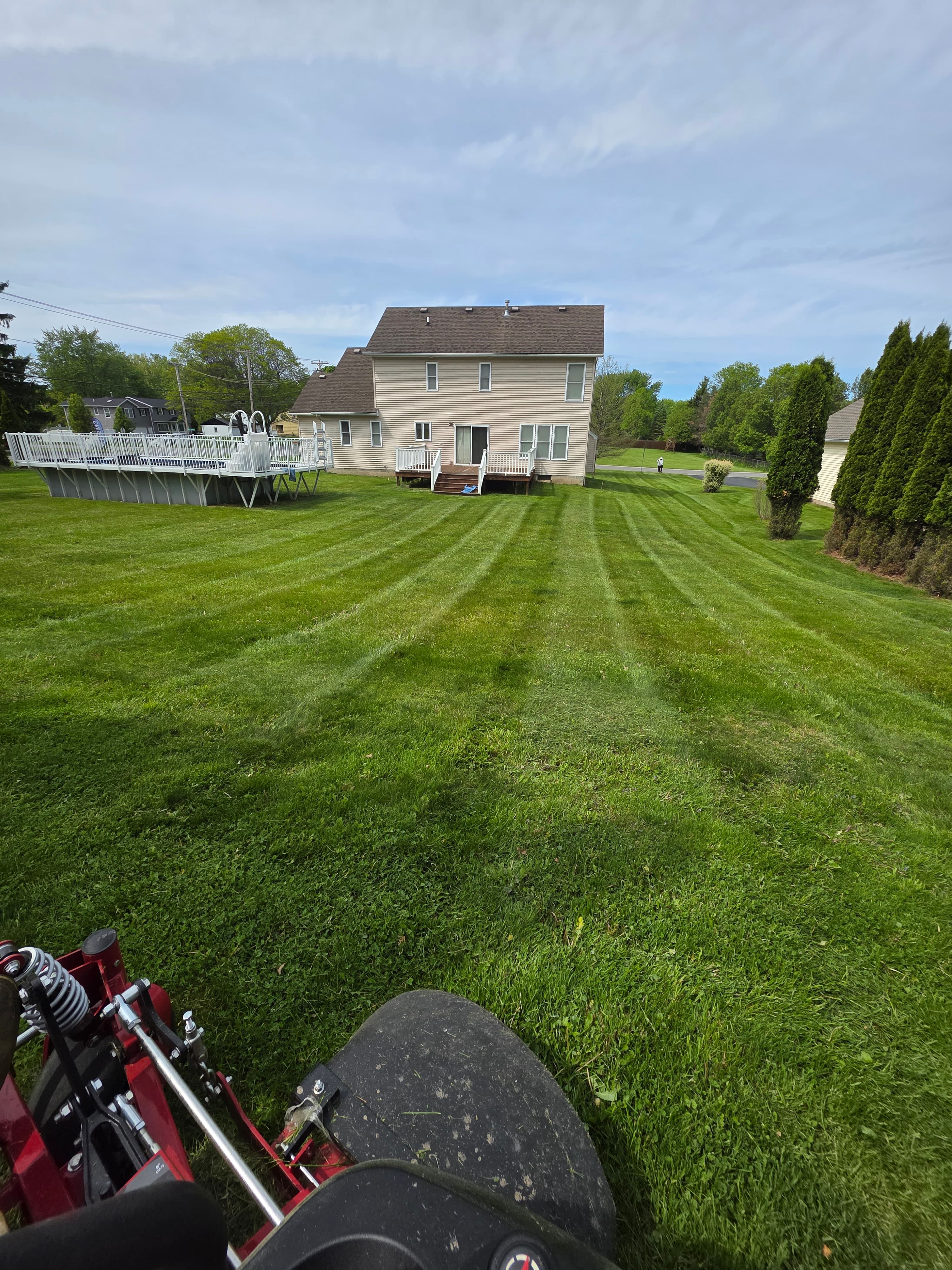 Striped green lawn behind a tan house and pool, viewed from a red lawnmower.